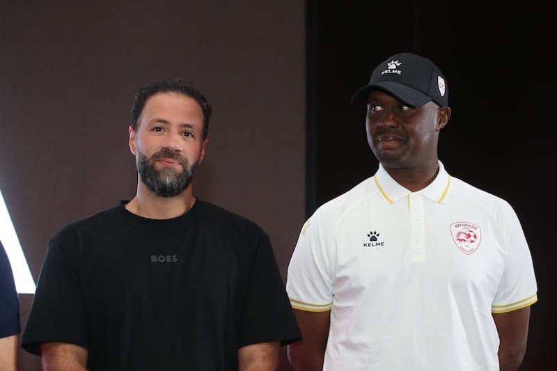 Two men stand side by side indoors; one wears a black t-shirt, the other a white polo with a cap, both looking at the camera. Two men stand side by side indoors; one wears a black t-shirt, the other a white polo with a cap, both looking at the camera.