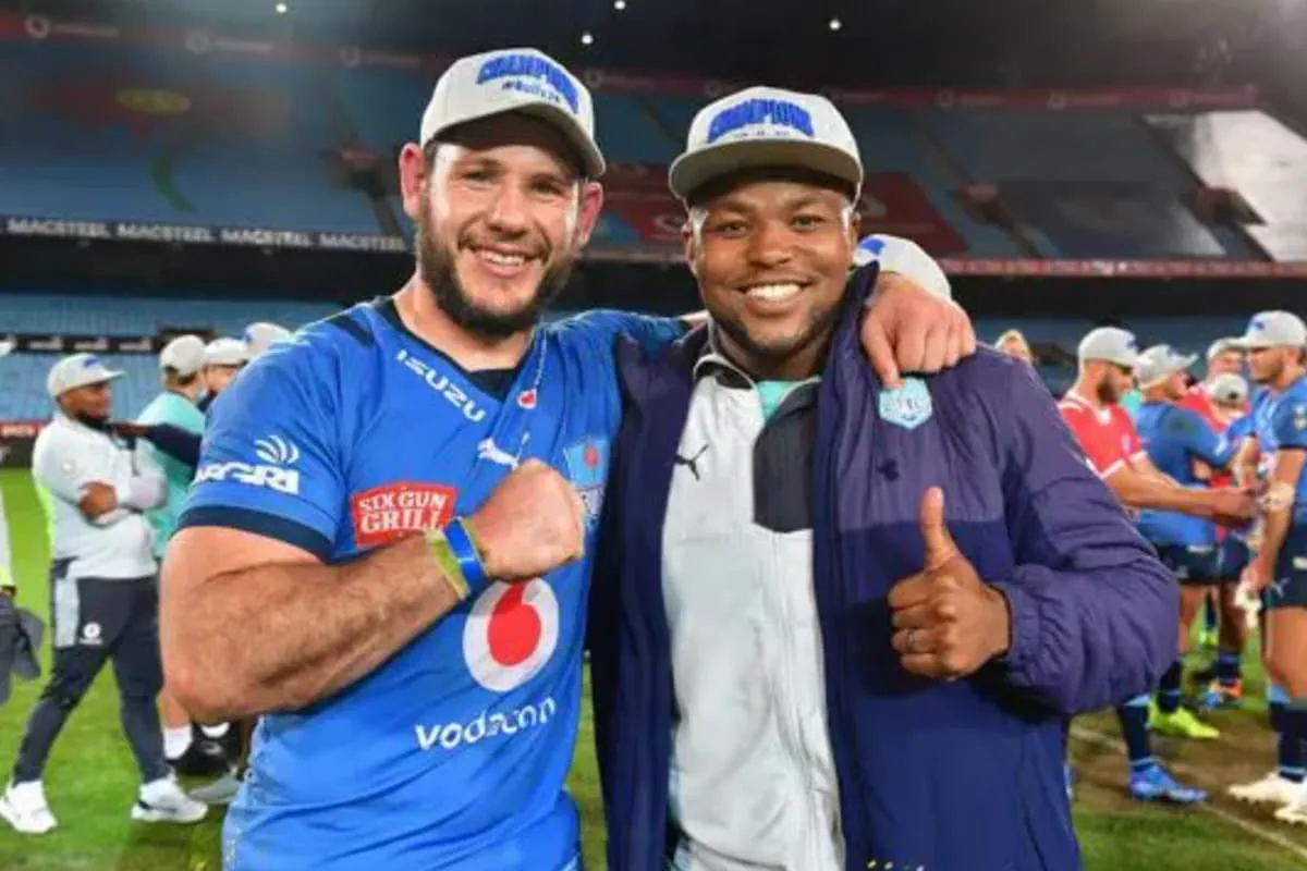 Two rugby players pose on the field, smiling with arms around each other; one wears a blue Vodafone kit, the other a jacket and cap.