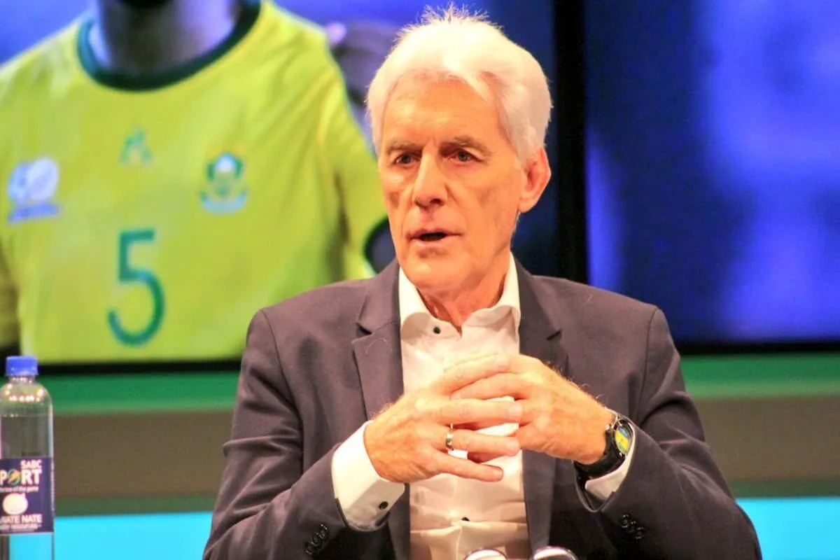 Older man in a dark suit and white shirt sits with hands clasped, speaking in a studio setting. Older man in a dark suit and white shirt sits with hands clasped, speaking in a studio setting.