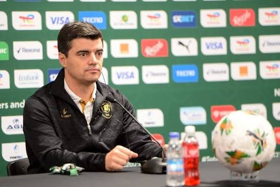 Man in a dark jacket sits at a press conference table with a microphone, a football, and bottled drinks in front, against a backdrop of sponsor logos.