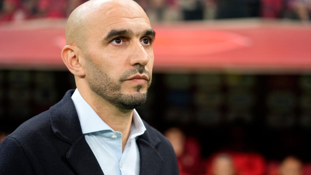 Man with a shaved head and trimmed beard wearing a dark coat and light blue shirt, standing in a stadium stands area. Man with a shaved head and trimmed beard wearing a dark coat and light blue shirt, standing in a stadium stands area.