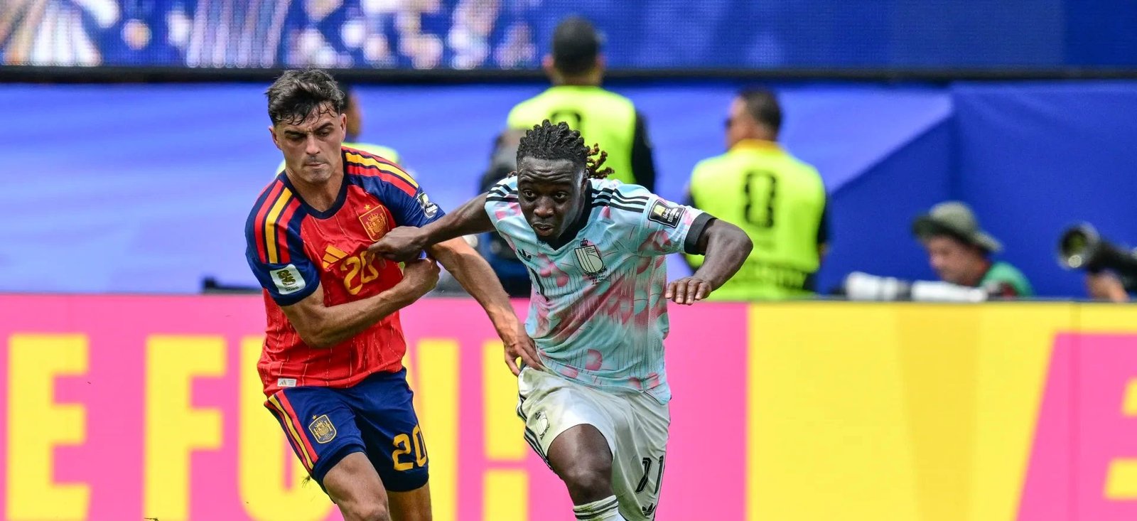 Two football players sprint for the ball during a match; one in red-blue kit, the other in a pale pink-and-white jersey. Two football players sprint for the ball during a match; one in red-blue kit, the other in a pale pink-and-white jersey.