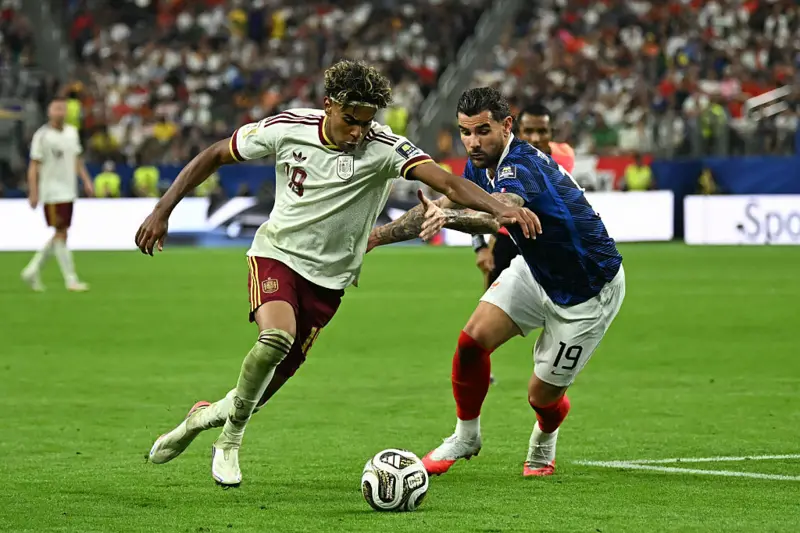 Two soccer players chase the ball on a green field during a competitive match; one wears a white jersey with number 8 and maroon shorts, the other a blue jersey with white shorts and red socks. Crowd visible in the background.