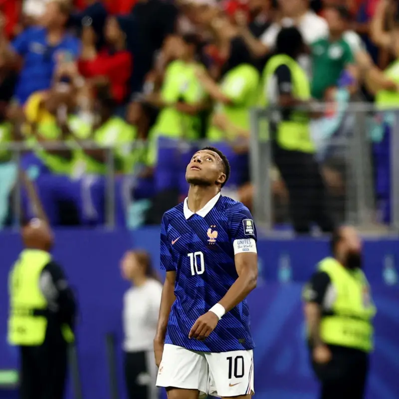 French national team footballer in blue jersey with number 10, looking up toward the sky on the pitch, with a blurred crowd in the stands behind him.