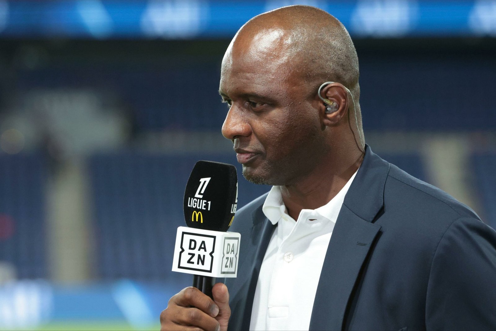 Male sports presenter with a wired in-ear monitor holding a DAZN microphone during a live interview.