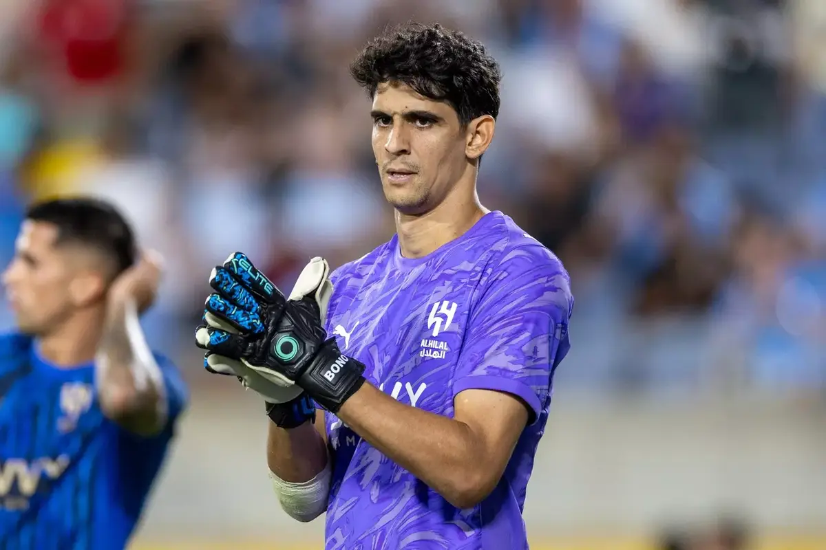 Goalkeeper in a purple jersey with patterned design, wearing gloves, standing on a football pitch with hands clasped together. Goalkeeper in a purple jersey with patterned design, wearing gloves, standing on a football pitch with hands clasped together.