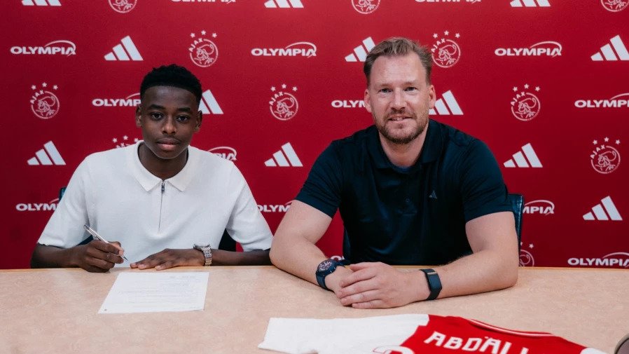 Young man signs a document at a table as a man in a dark polo sits beside him, with a red branded backdrop behind them.
