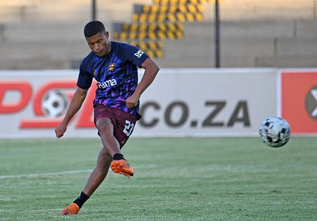 Male soccer player in blue jersey and maroon shorts kicking a ball on a green field during a match, orange cleats visible.
