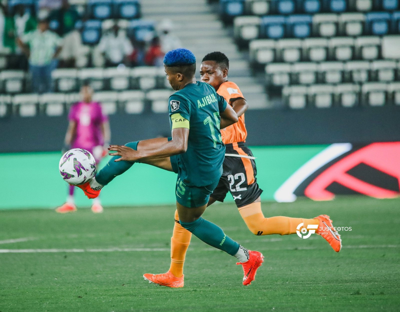 Two soccer players compete for the ball on a green pitch, one in teal with blue hair and captain armband, the other in orange, stadium seats blurred in the background.