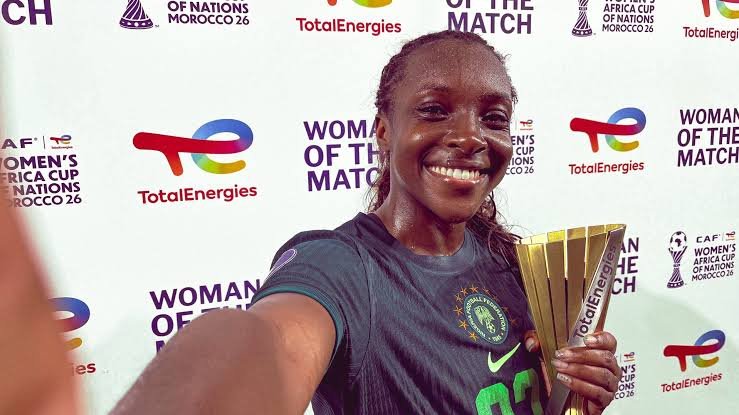 Smiling female footballer in a green jersey holds a gold trophy at a 'Women of the Match' event backdrop.