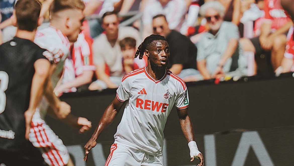 Soccer player with braided hair in a white jersey marked REWE, running on the pitch with fans in the stands behind.