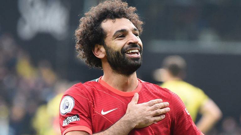 Soccer player in a red jersey smiles with his right hand on his chest during a match on the field.