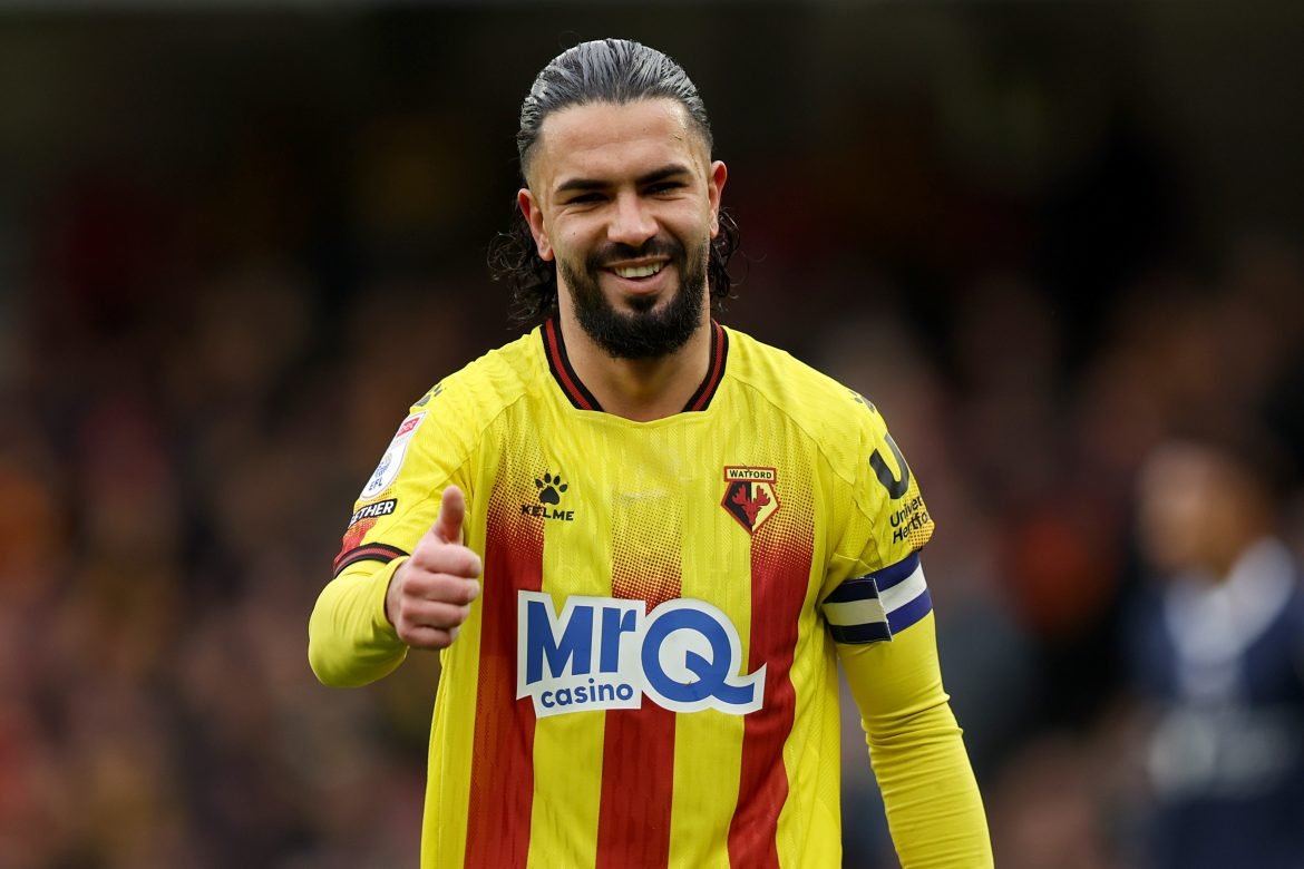 Watford FC player in a yellow and red striped jersey giving a thumbs up on the pitch, with the MRQ casino sponsor visible on the front.