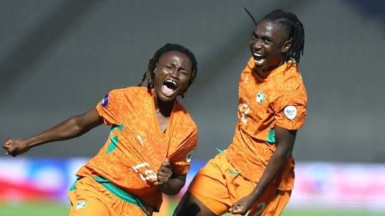 Two football players in orange uniforms celebrate on the field, one shouting with clenched fists as the other smiles nearby.