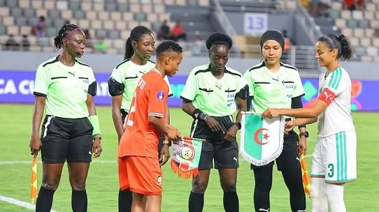 Female referees in neon green uniforms oversee a coin toss on a soccer field as captains from orange and white/green teams exchange banners before the match.