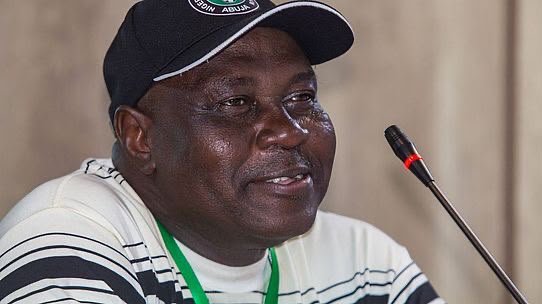 Man wearing a black cap and striped shirt speaks into a microphone at a conference panel.