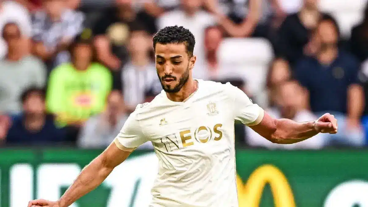 Soccer player in a white kit with the EOS sponsor, in mid-action on the field with a blurred crowd behind him. Soccer player in a white kit with the EOS sponsor, in mid-action on the field with a blurred crowd behind him.