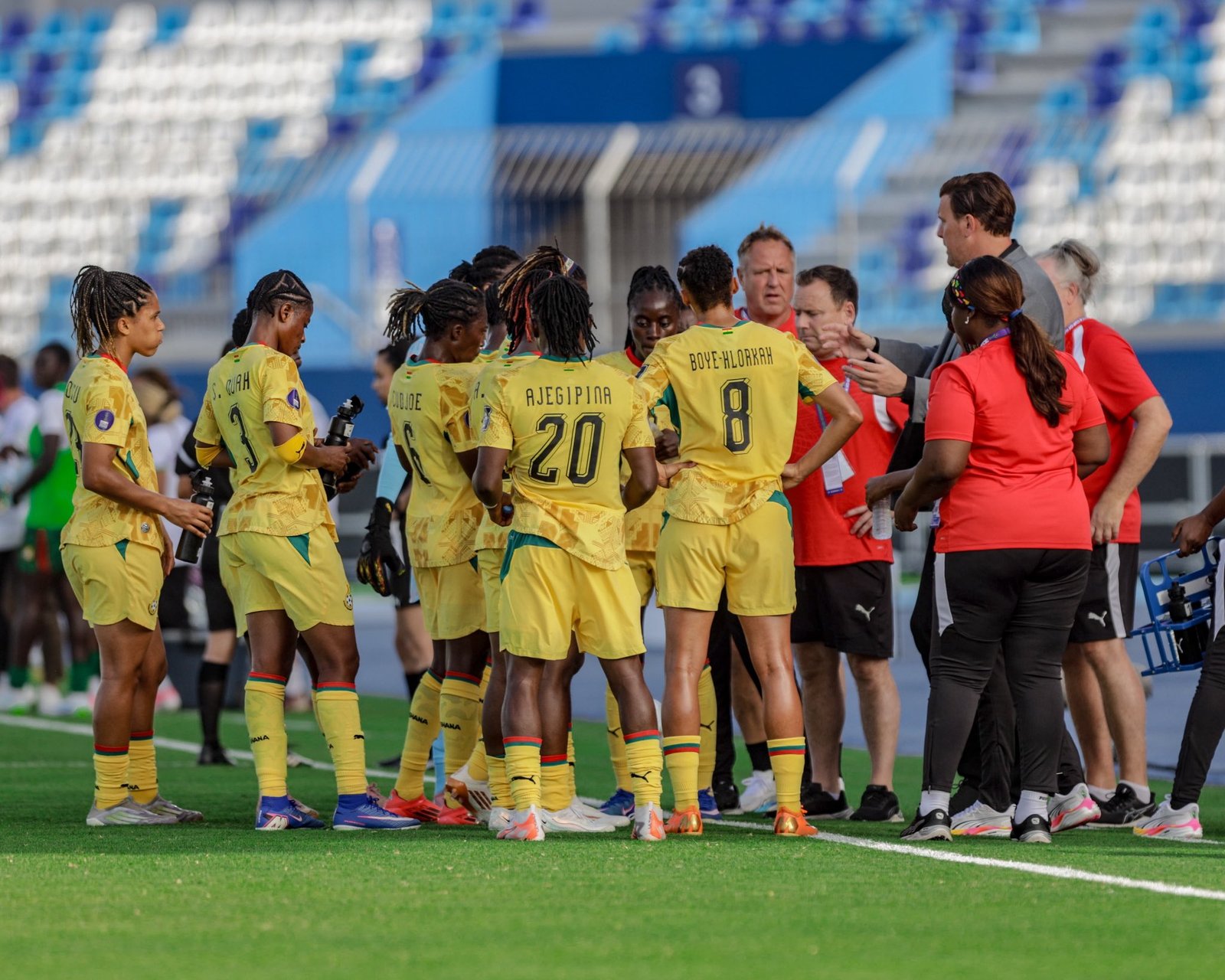 Group of female soccer players in yellow uniforms huddle on a green field while coaches in red shirts guide them.