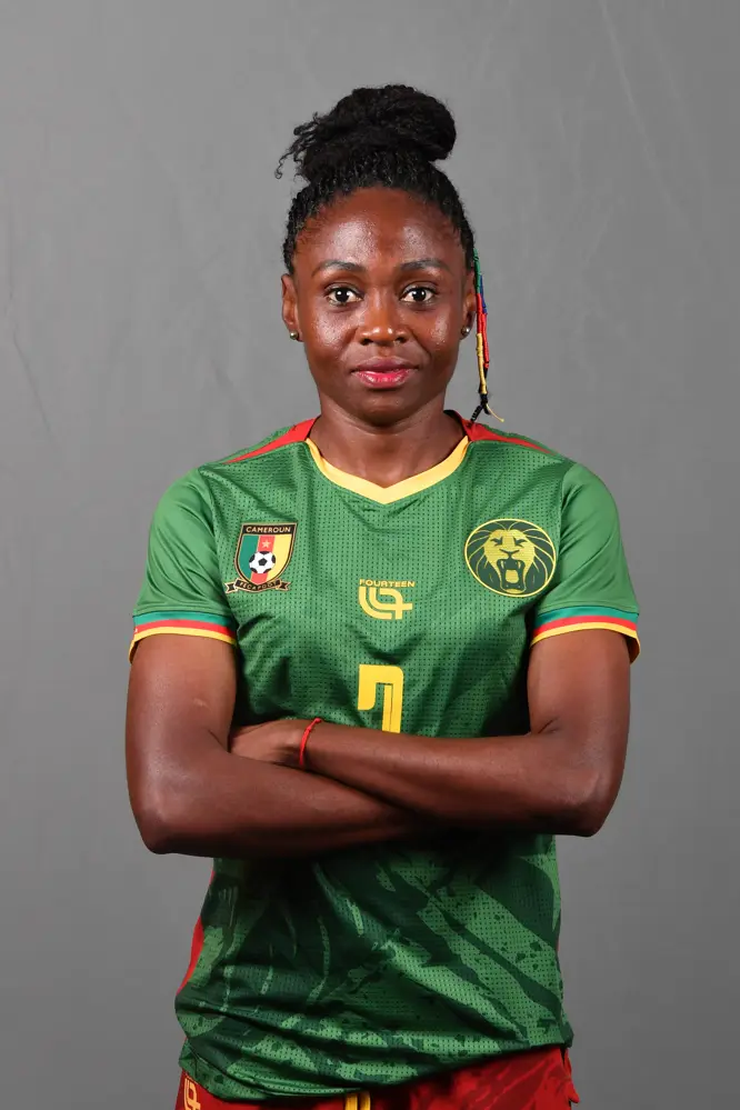 Female Cameroonian footballer wearing the national team green jersey, arms crossed, facing the camera.