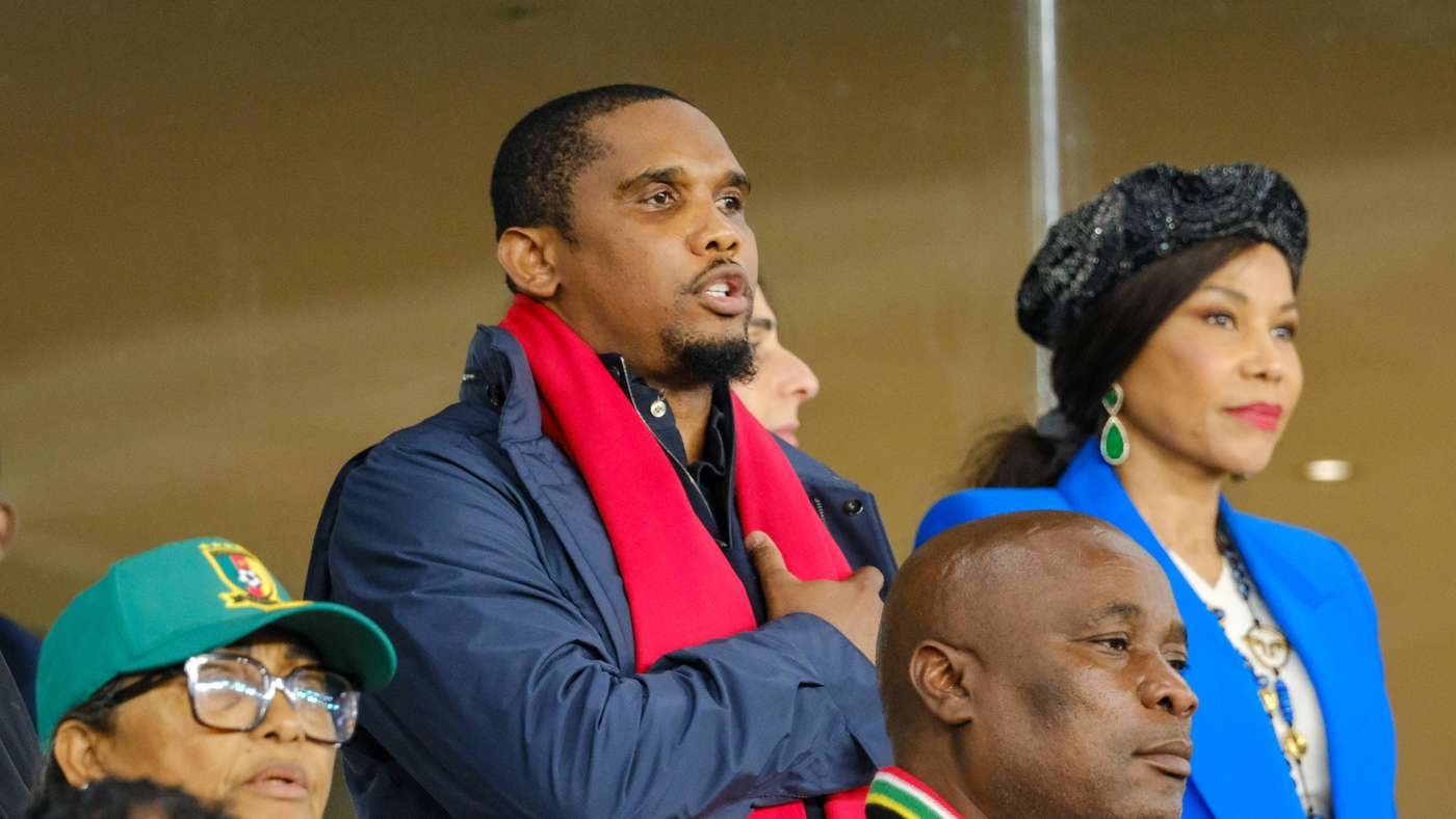 Man in navy jacket with a red scarf speaking or singing, flanked by a woman in a bright blue blazer and decorative hat at an event. Man in navy jacket with a red scarf speaking or singing, flanked by a woman in a bright blue blazer and decorative hat at an event.