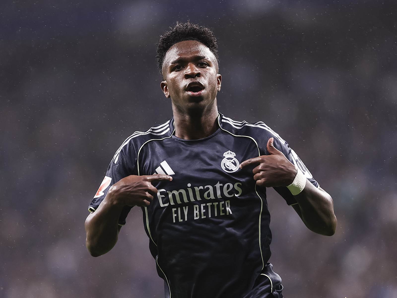 Soccer player in a dark Real Madrid jersey celebrating, hands on his chest near the crest, rain in the background