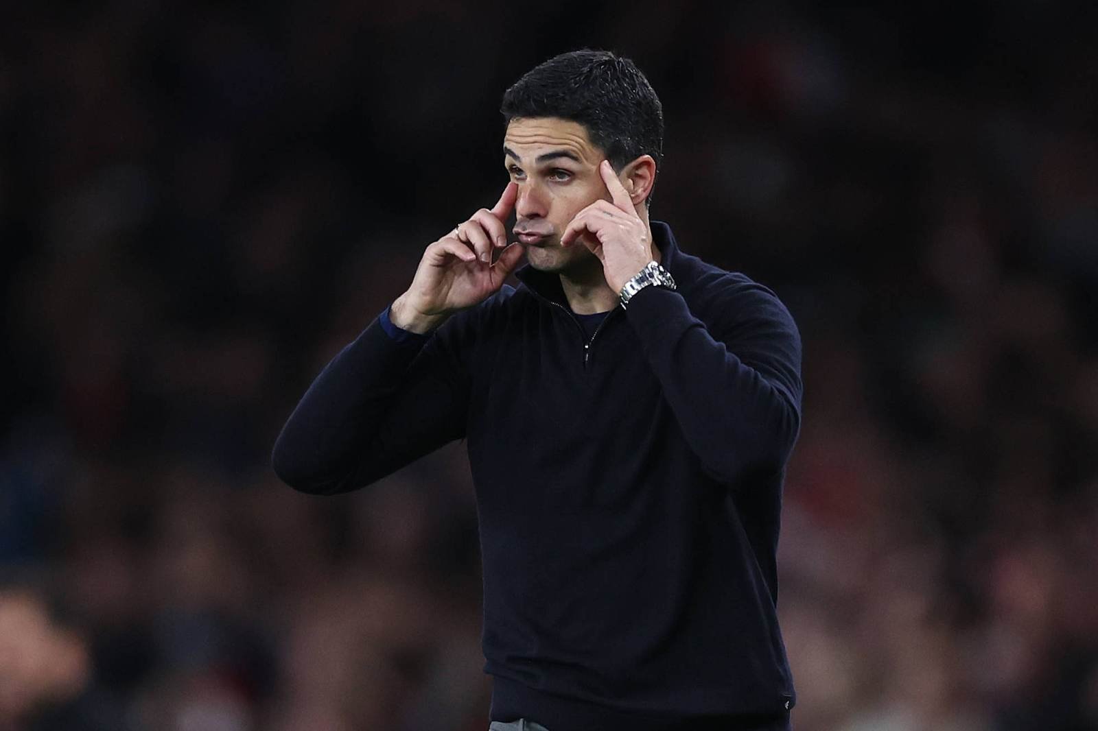 Man on a sports field wearing a dark sweater, pressing his index fingers to his temples with a pensive look.