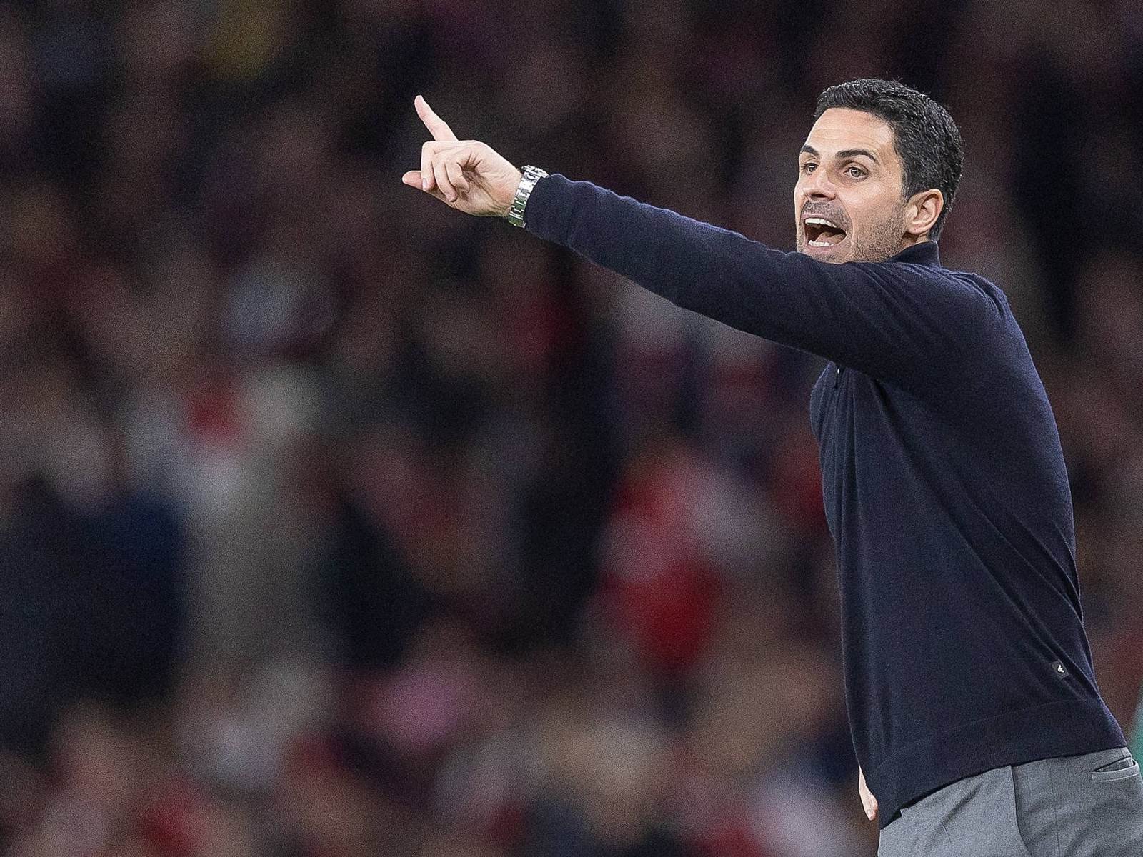 Coach gesturing with a raised arm and pointing finger during a game, blurred stadium spectators in the background