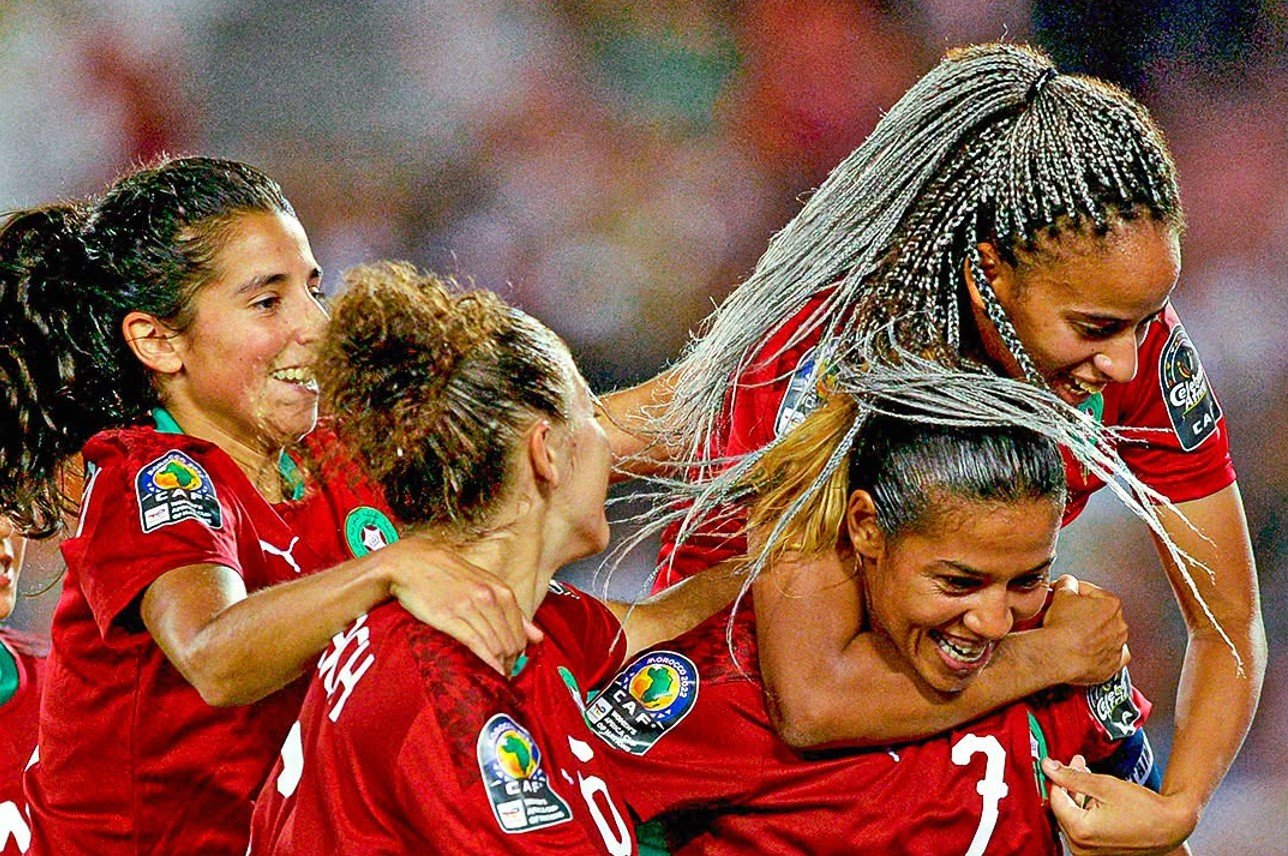Women’s soccer team celebrates a goal, huddled together in red uniforms with Africa Cup of Nations patches.