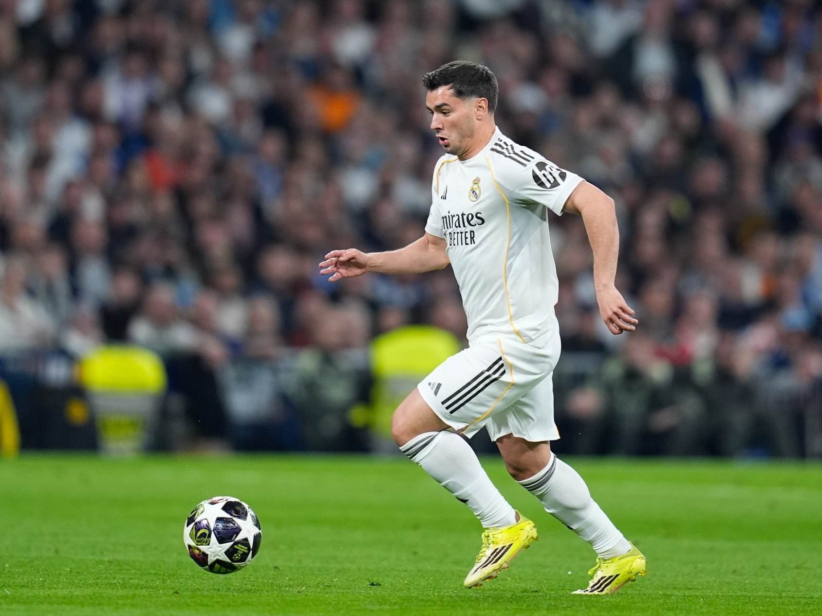 Soccer player in white Real Madrid kit dribbling a ball on a green pitch during a match with a crowded stands backdrop.