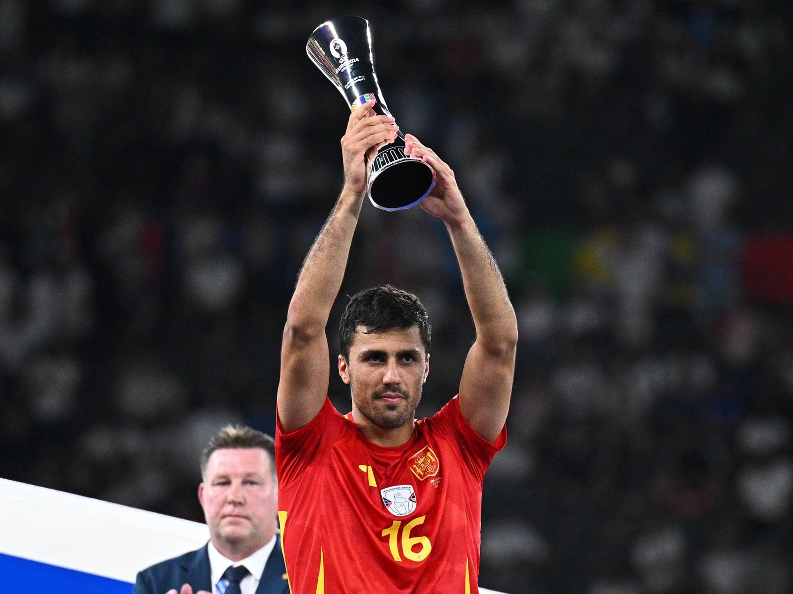 Soccer player in a red jersey lifts a silver trophy overhead to celebrate victory on the field.