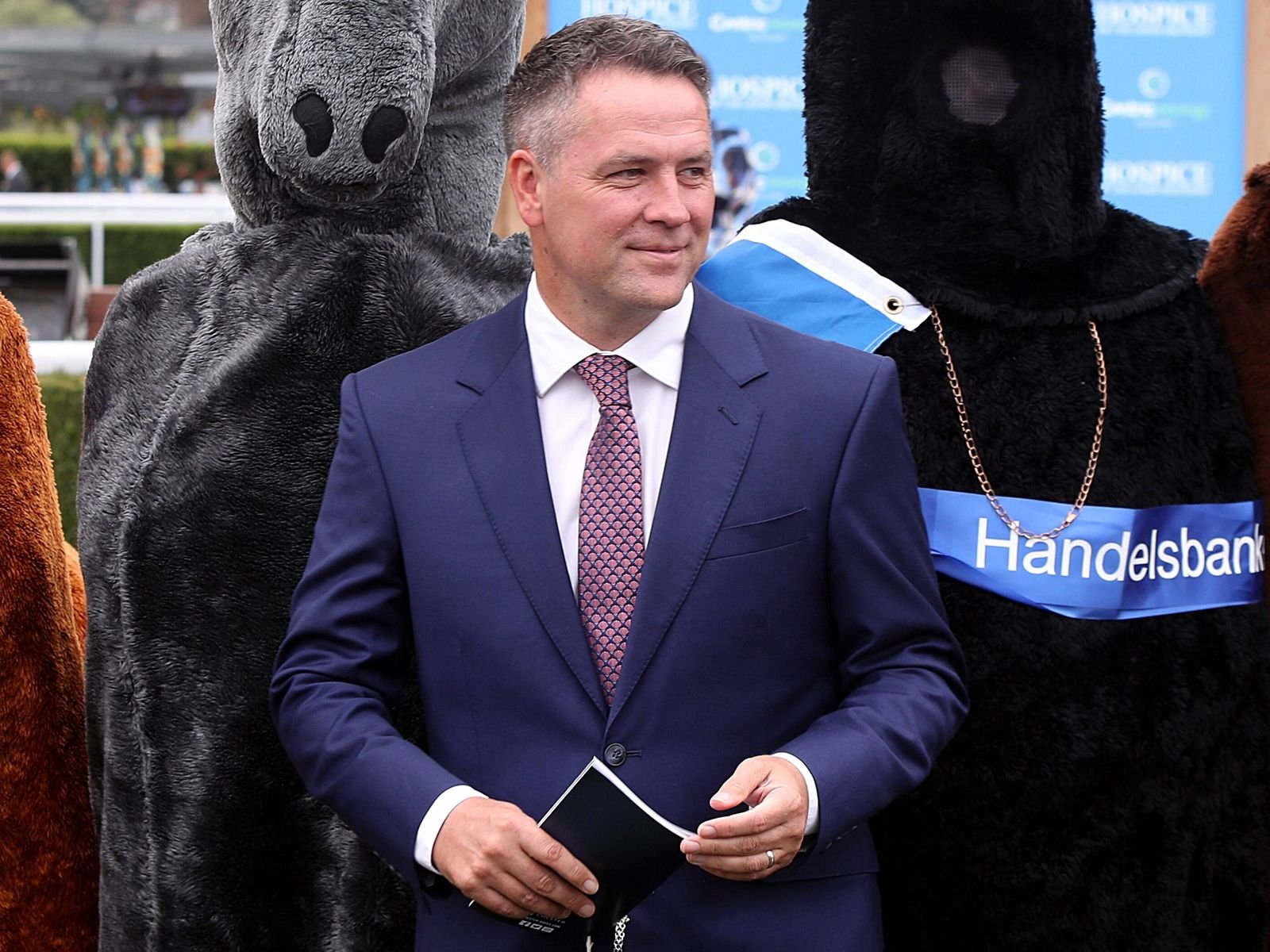 Man in a blue suit and pink patterned tie holds a booklet, flanked by large gray and black bear mascots at a Handelsbank event.