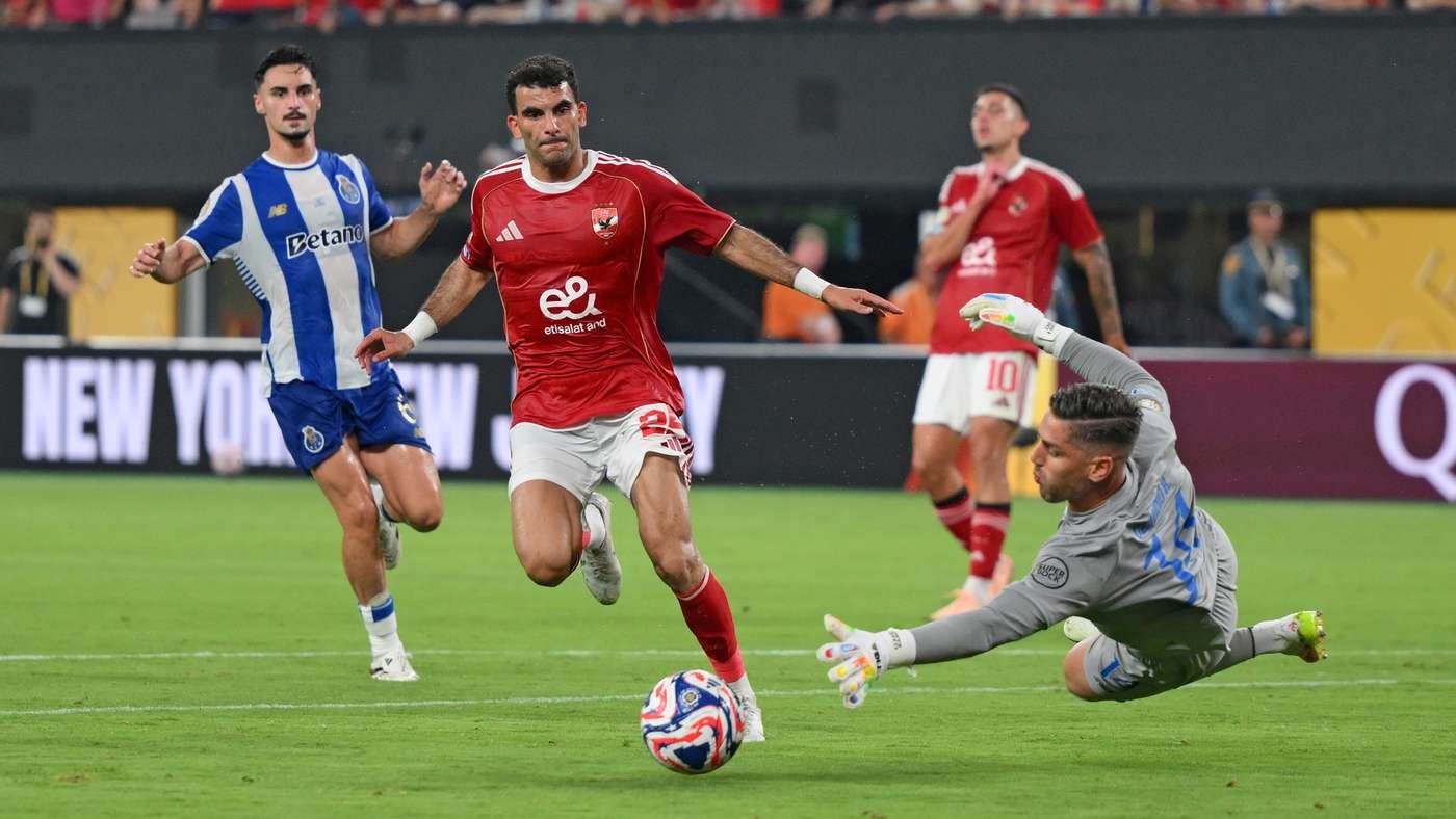 Red-jersey attacker dribbles toward the ball as a goalkeeper dives to make a save in a live football match.