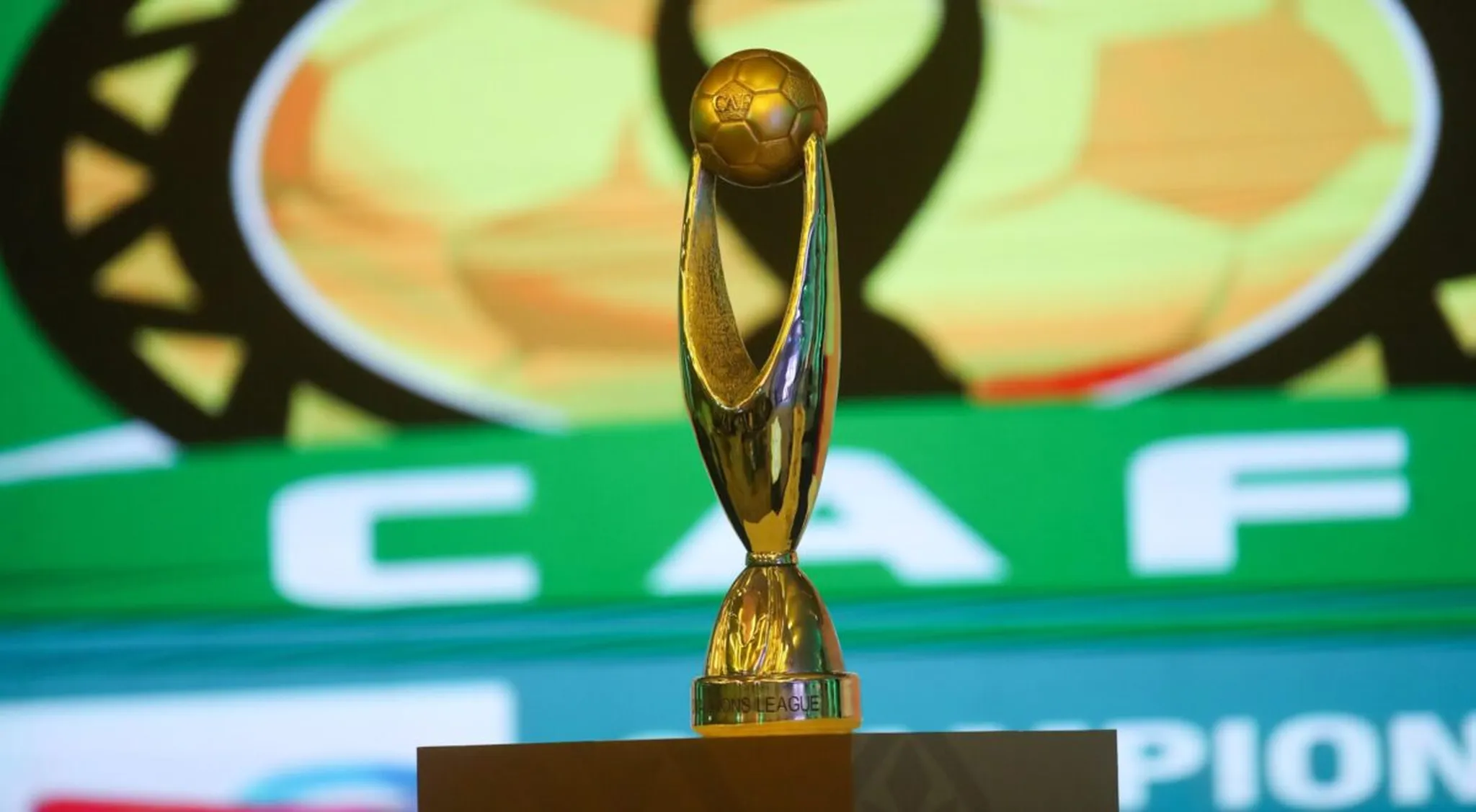 Gold trophy on a pedestal during an awards ceremony with a blurred green banner in the background, centered in the image.