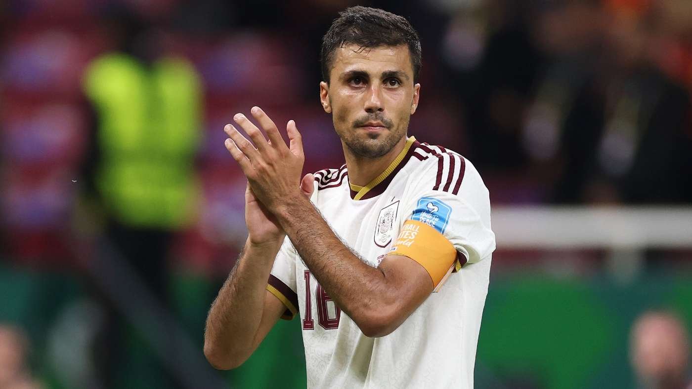 Male soccer player in a white maroon-trim kit clapping on the field, wearing an orange captain's armband on his left arm.