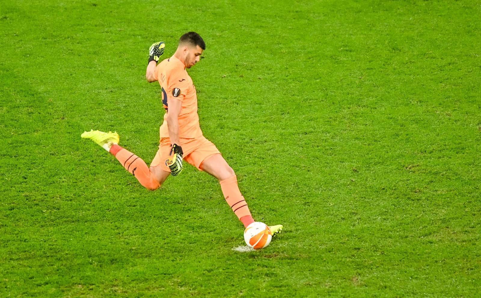 Soccer goalkeeper in orange kit kicking an orange–white ball on a green pitch with gloves on