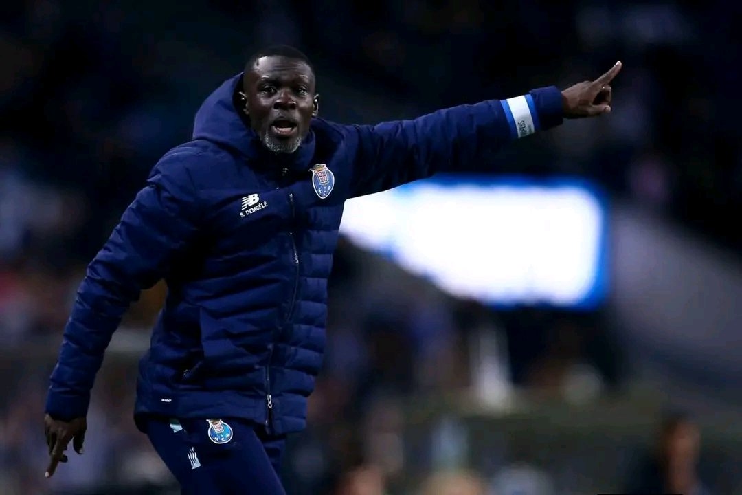 Football coach on the sideline in a dark blue padded jacket, pointing emphatically with his right arm during a match.