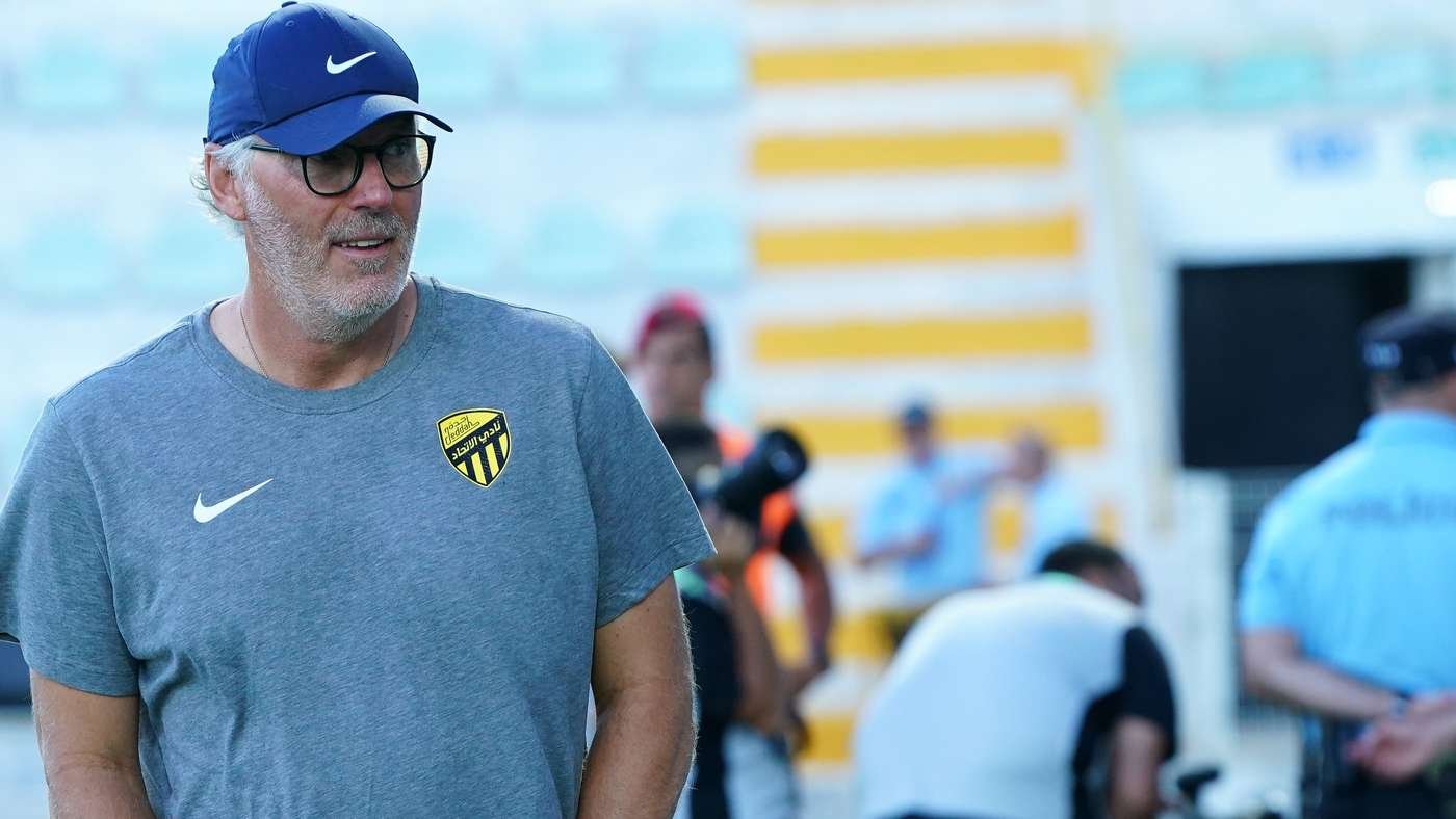Man wearing a blue Nike cap and gray shirt with a yellow-and-black crest, standing outdoors and looking to the left.