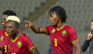 Three footballers in red Cameroon jerseys on the pitch, one pointing as teammates look on in the stands. Three footballers in red Cameroon jerseys on the pitch, one pointing as teammates look on in the stands.
