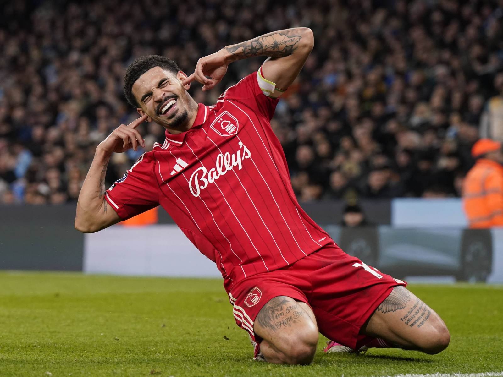 Footballer in a red striped kit celebrating a goal, kneeling on the grass with fingers to his ears as crowd cheers in the background.
