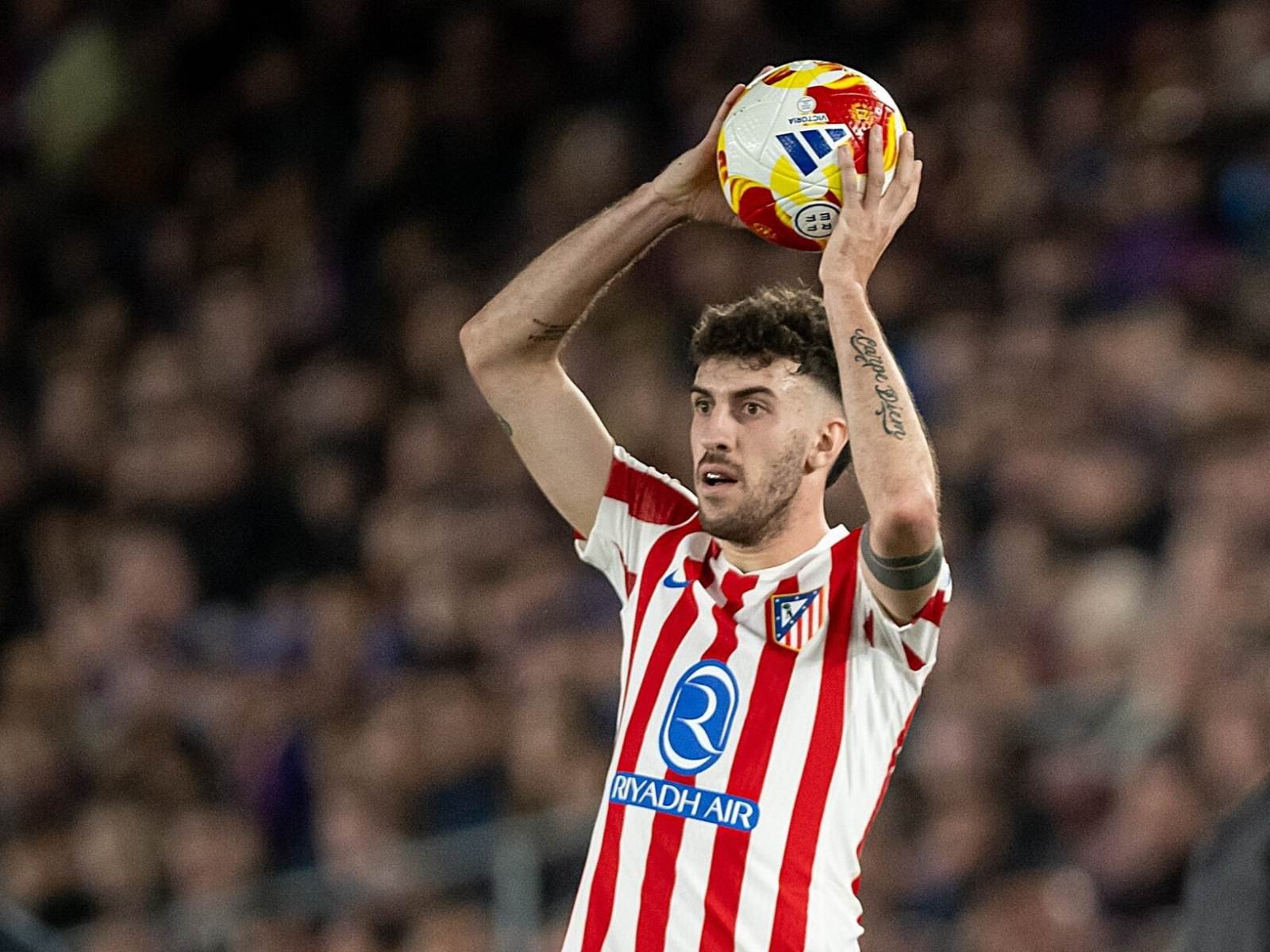 Soccer player in red-and-white striped jersey raises a colorful ball above his head during a match, crowd blurred in background