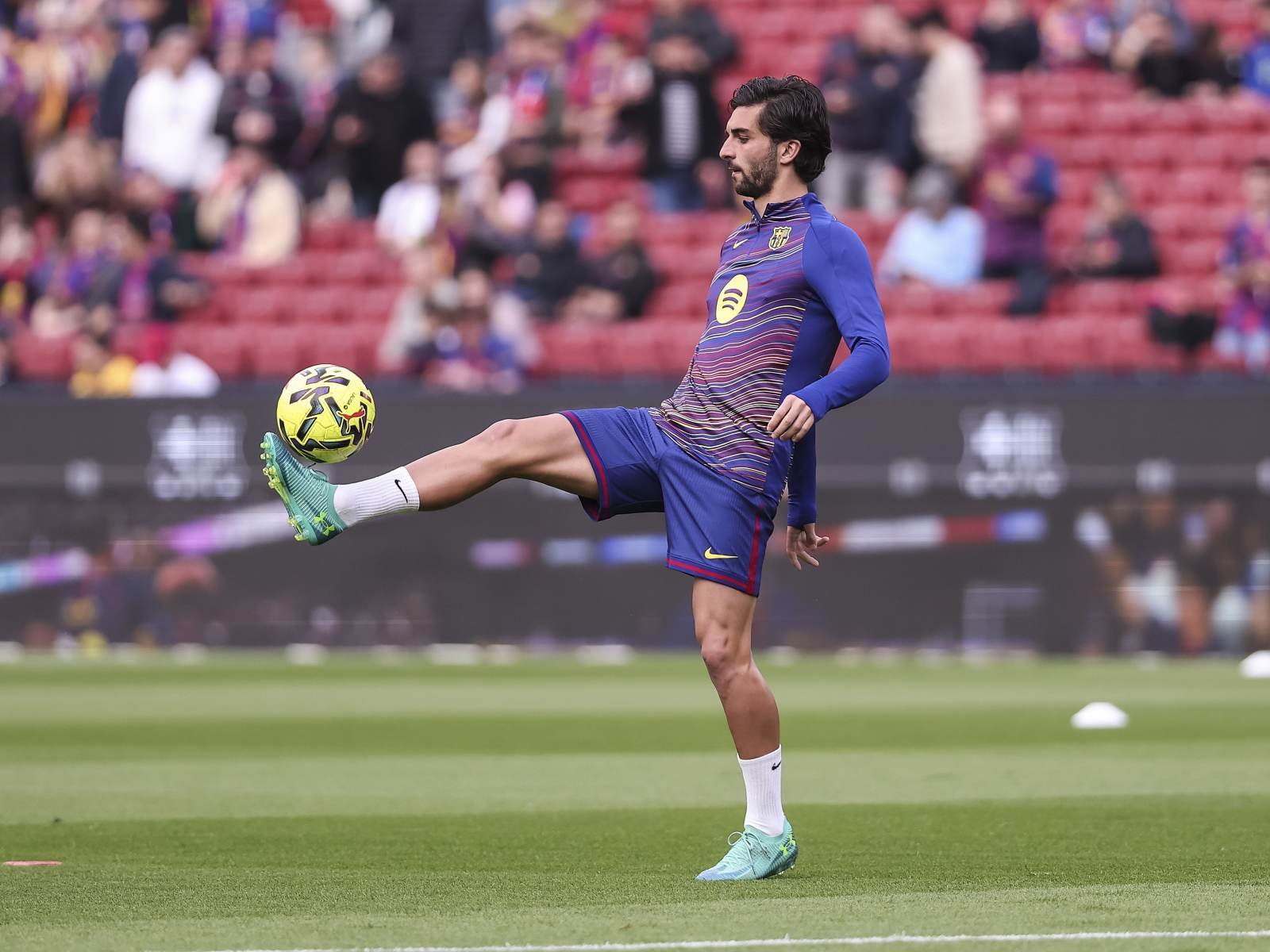 Soccer player in blue Barcelona kit traps a ball mid-air on a green field, crowd in the stands behind.