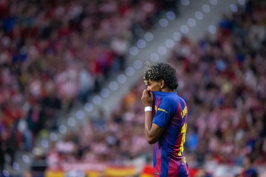 Soccer player in Barcelona blue-and-maroon kit covers mouth with his hand on a crowded stadium background