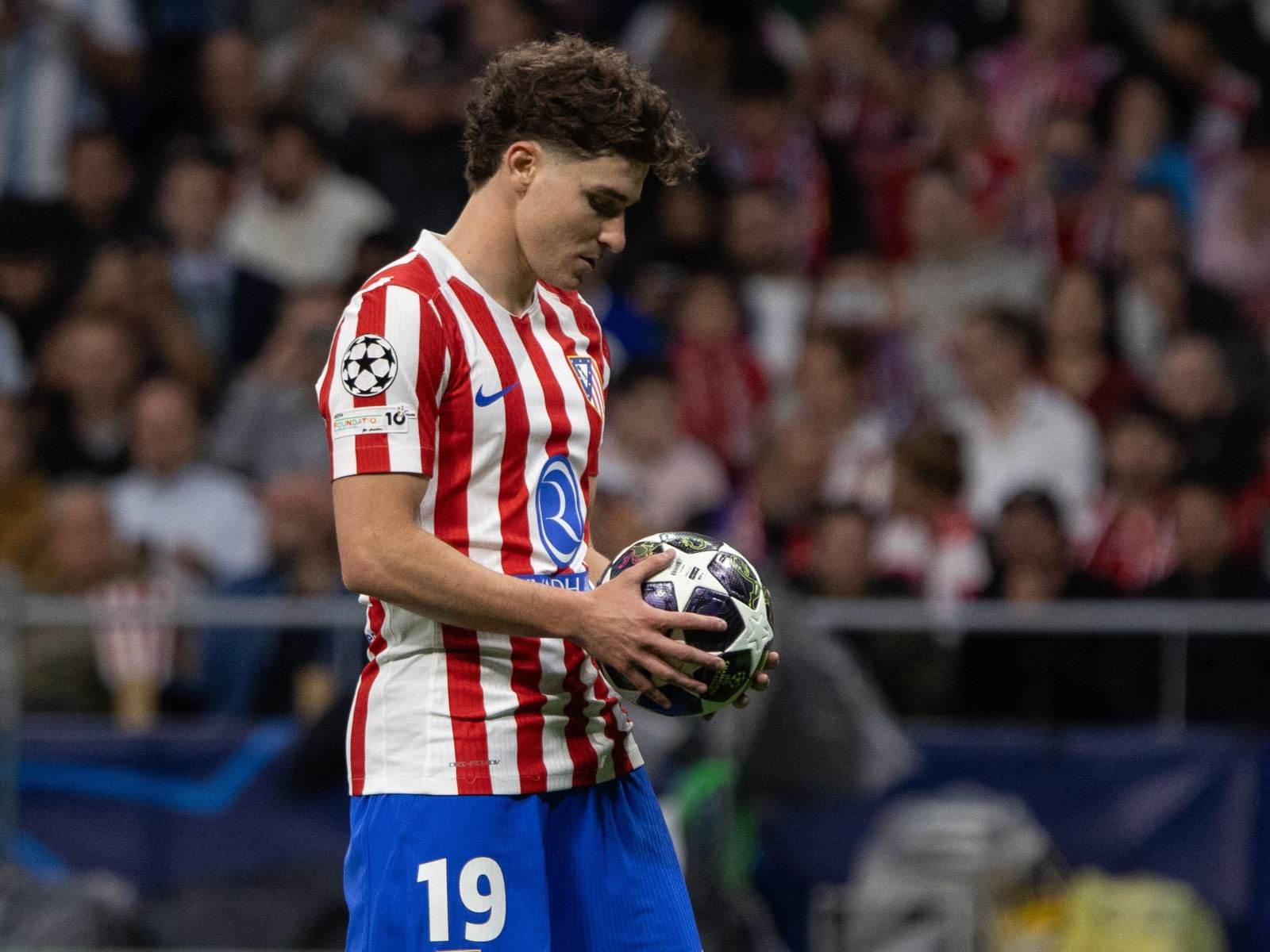 Soccer player in a red-and-white striped jersey and blue shorts holding a ball on the field, with a crowded stadium in the background.