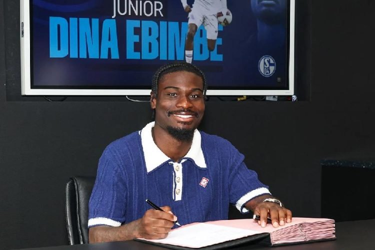 Man in a blue polo shirt signs a document at a desk, smiling, with a stack of pink folders nearby and a screen behind displaying 'DINA EBINDE'.