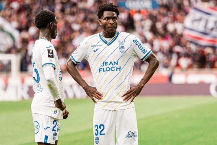 Two footballers in white kits with blue-yellow diagonal stripes stand on a grassy pitch during a match, one facing the camera with hands on hips.