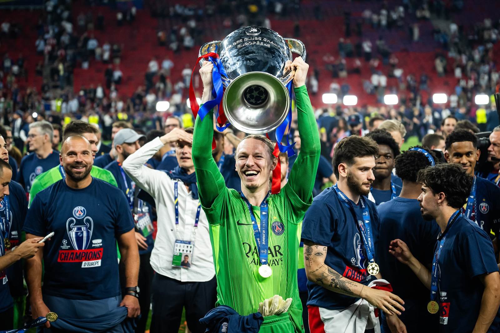 Goalkeeper in a green jersey lifts a large silver trophy above his head, celebrating with teammates on a stadium field after winning the Champions League.