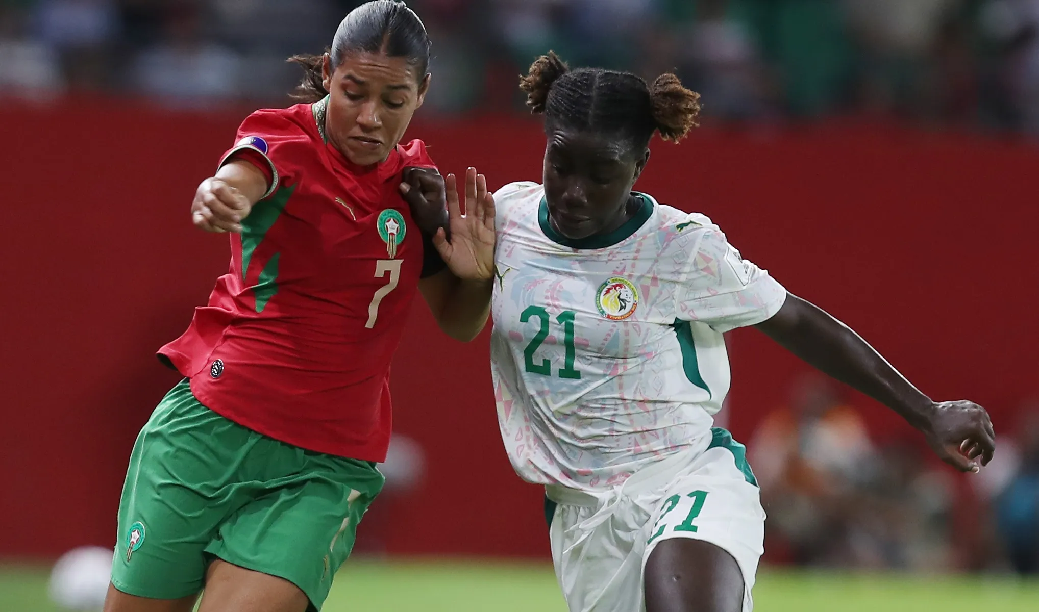 Two female soccer players clash for the ball in a high-action moment, one in red and green and the other in white and green uniforms on a green field.