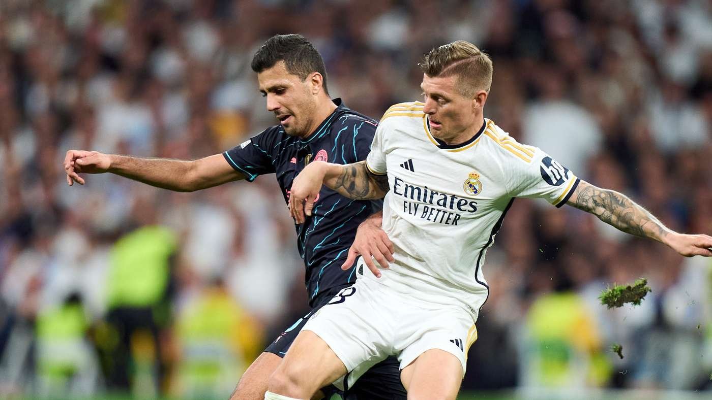 Two soccer players battle for the ball during a match, one in a white Real Madrid kit and the other in dark kit, with a blurred crowd in the background.