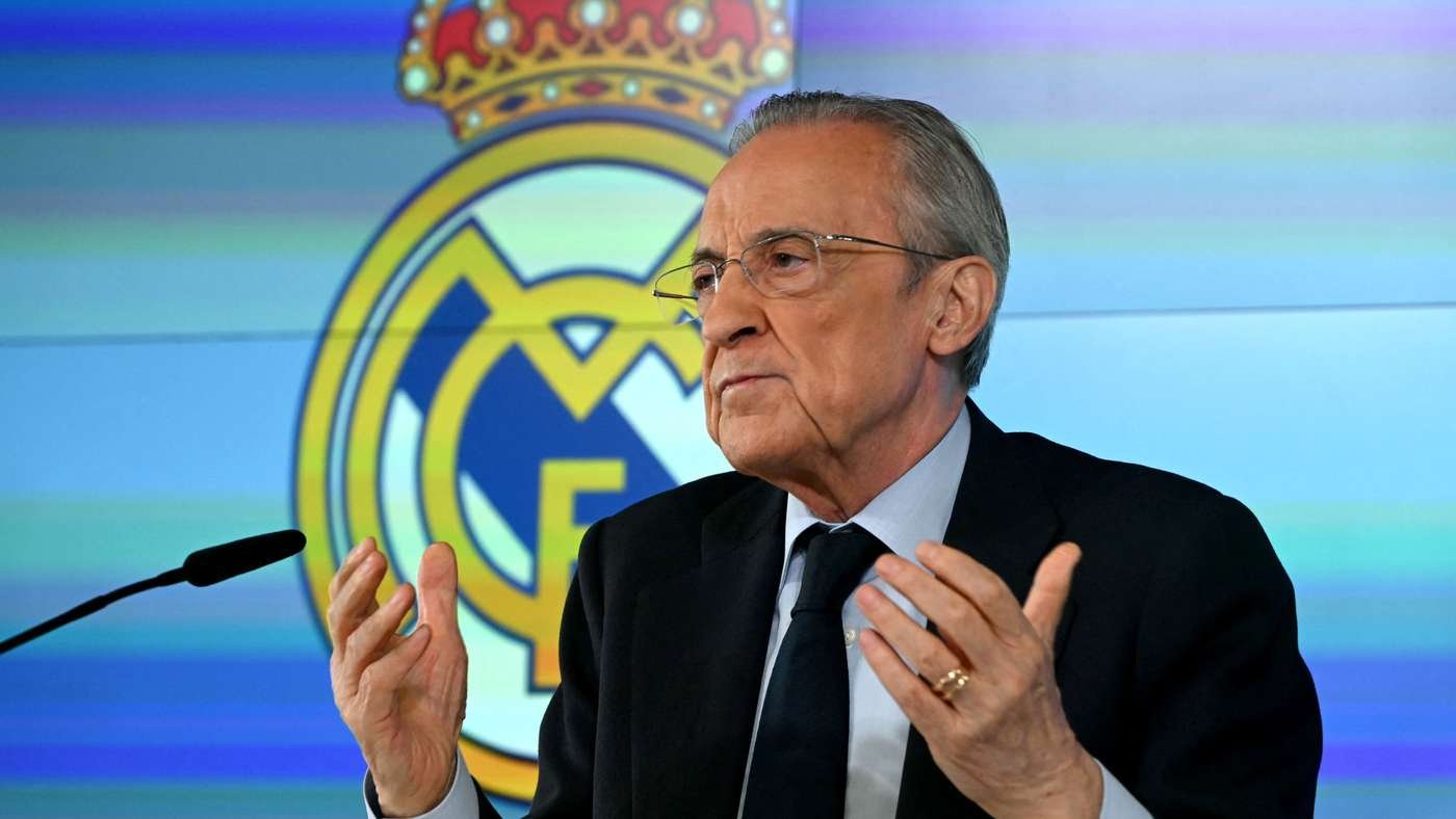 Older man in a dark suit speaks at a microphone with his hands raised; Real Madrid crest in the colorful background behind him.