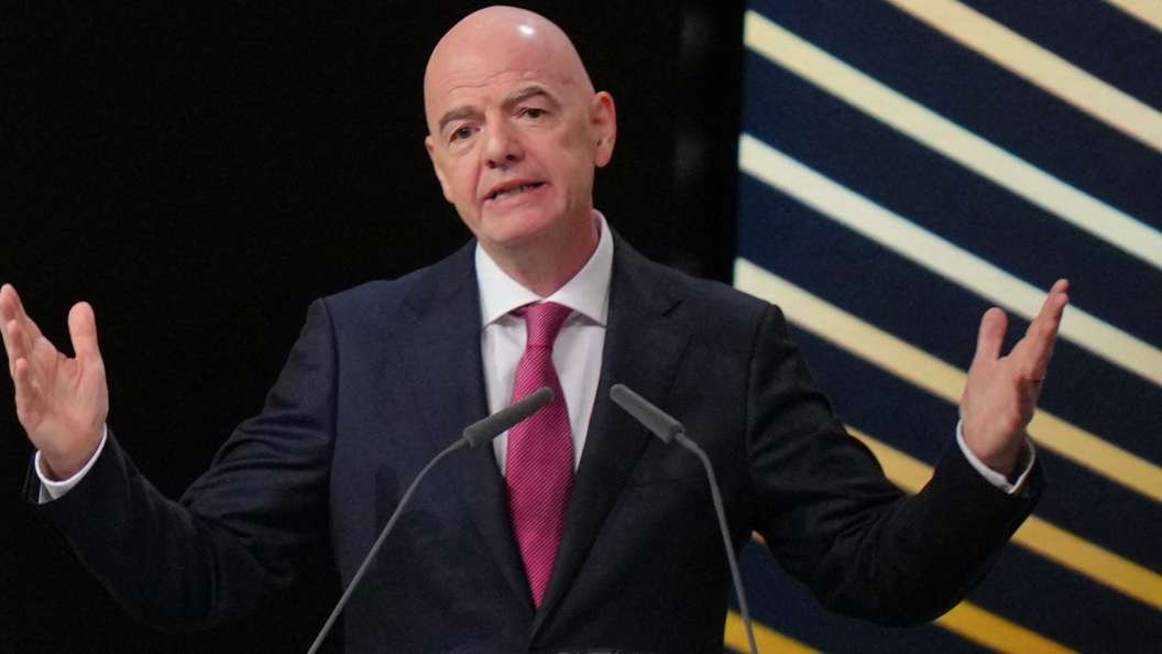 Bald man in a dark suit and red tie speaks from a podium with two microphones, gesturing with open hands.